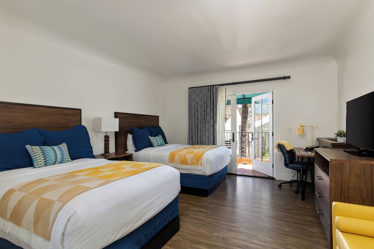 Quarto de hotel com duas camas, uma escrivaninha, uma cadeira, televisão e varanda com cortinas.