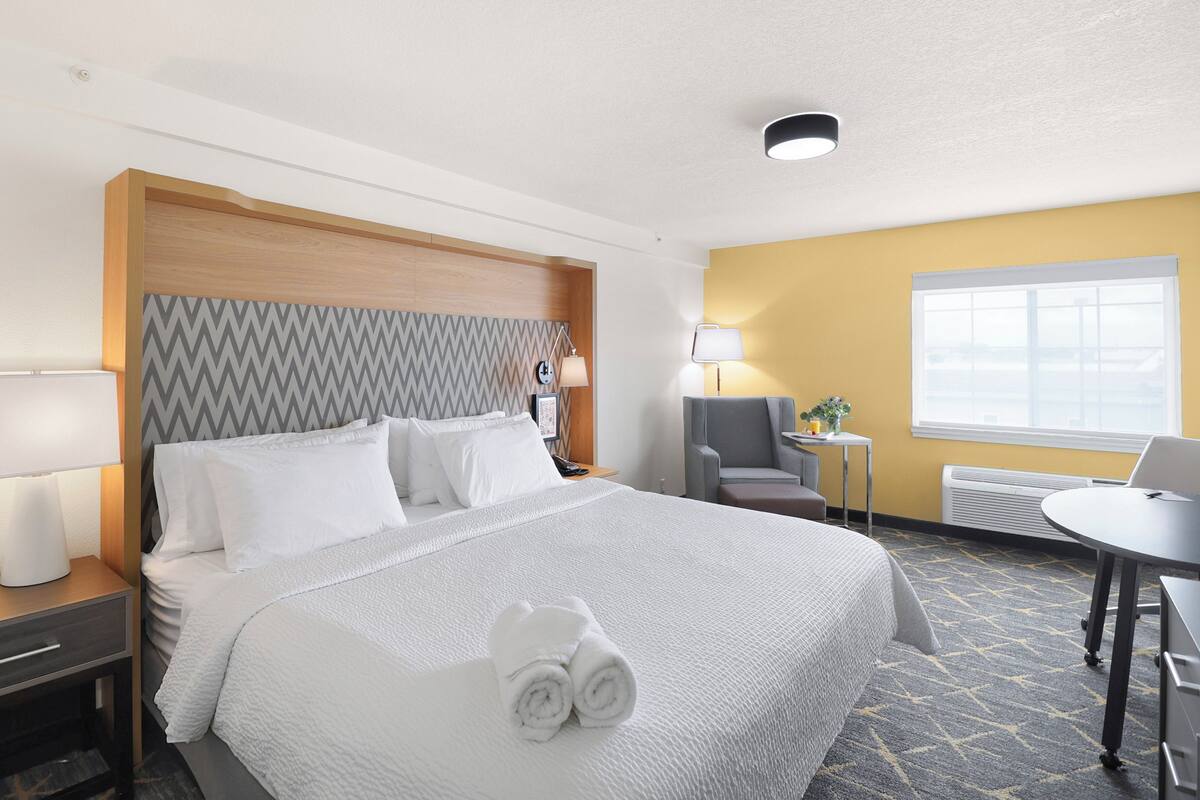 A hotel room with a bed, a desk, a chair, a lamp, and a window.