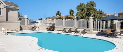 A swimming pool with lounge chairs and umbrellas, surrounded by a white fence and a building.