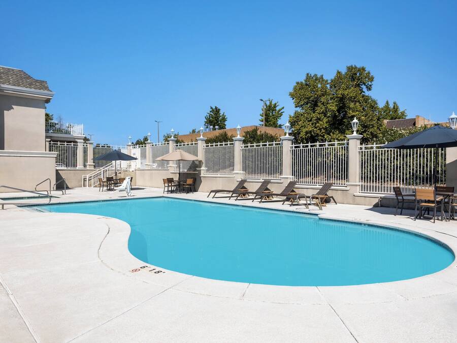 A swimming pool with lounge chairs and umbrellas, surrounded by a white fence and a building.