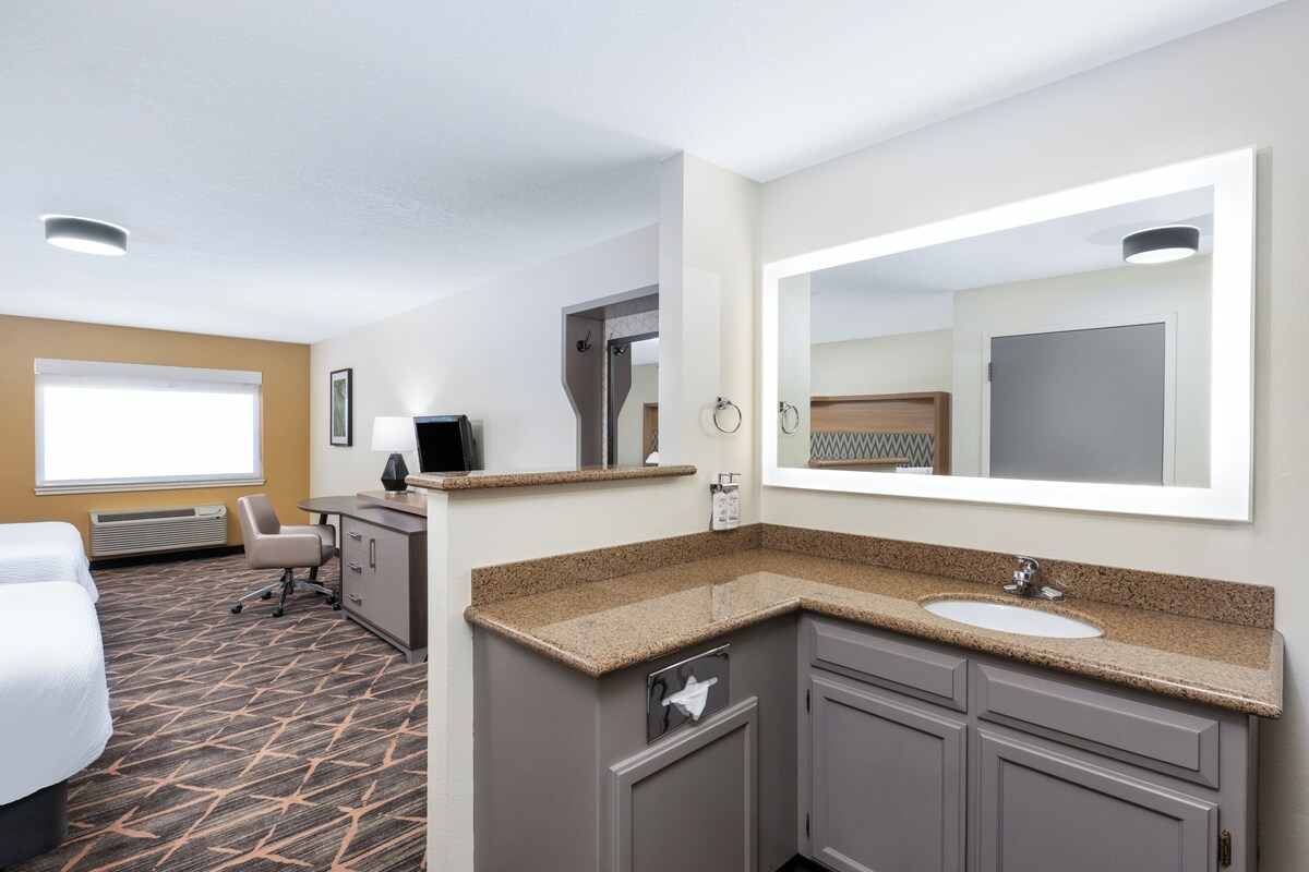 Habitación de hotel con cama, escritorio, silla, baño con lavamanos y espejo, y ventana con persianas.