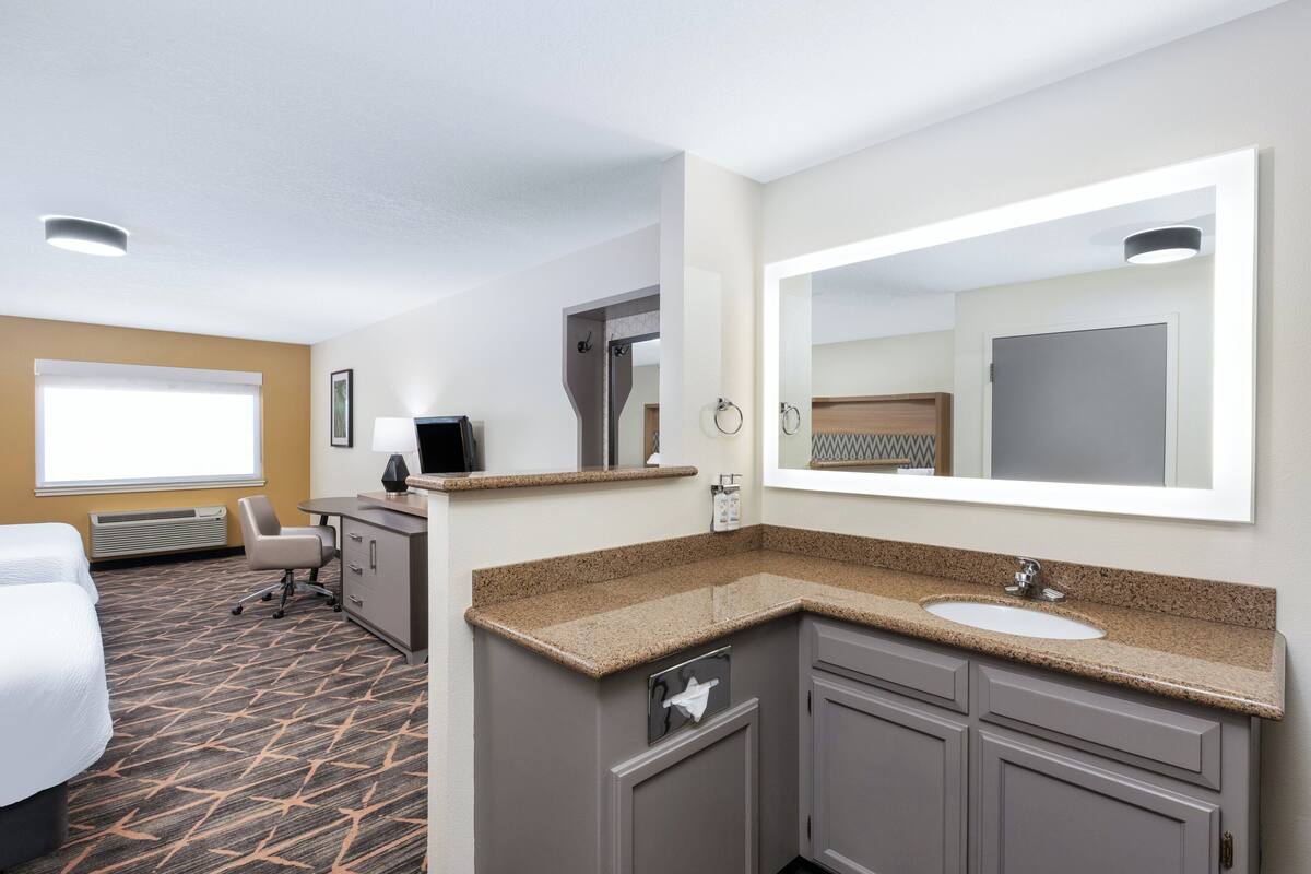 A hotel room with a bed, a desk, a chair, a bathroom with a sink and mirror, and a window.