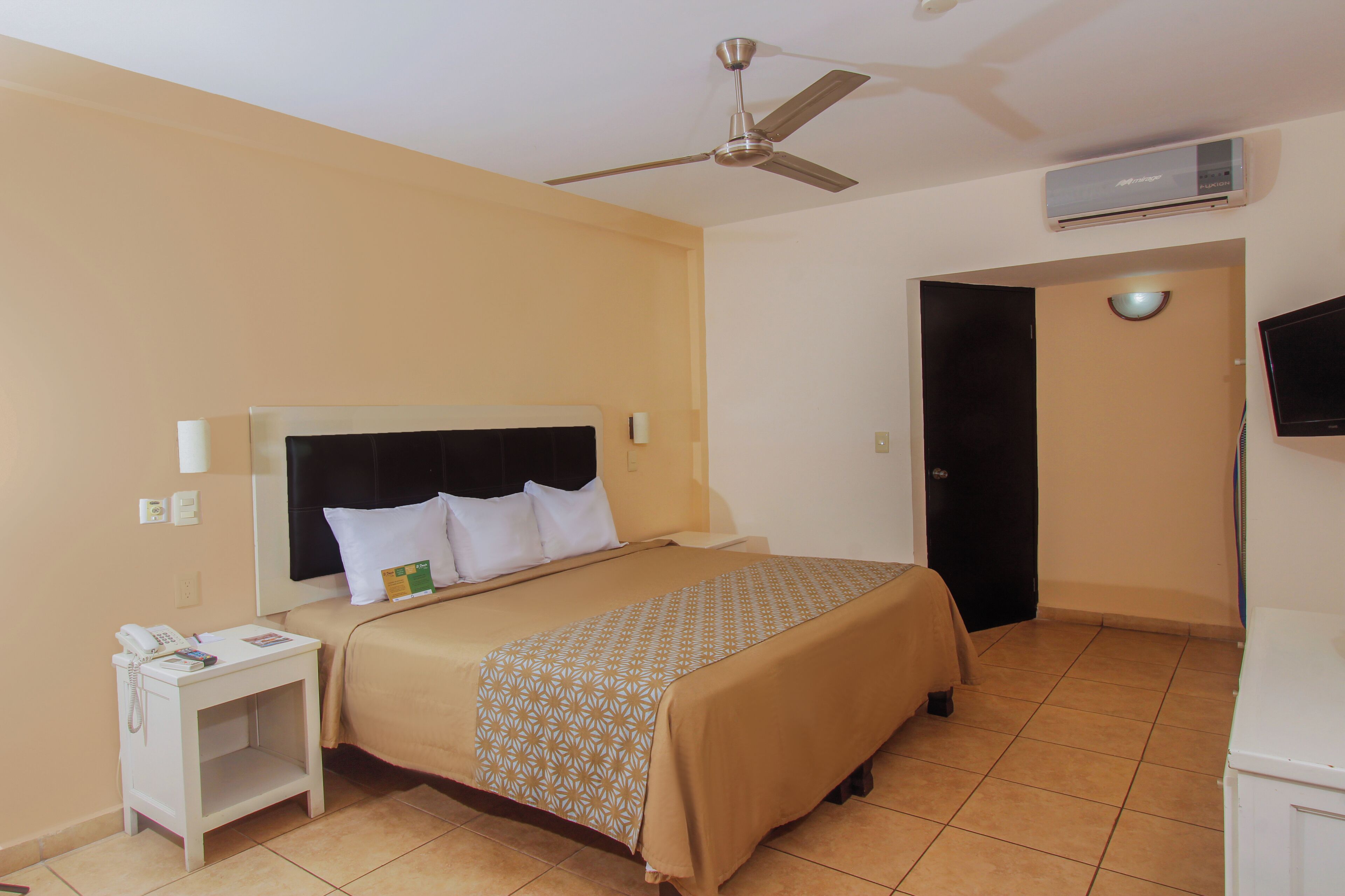 Una camera d'albergo con un letto, un comodino, una ventola a soffitto, una TV e un condizionatore d'aria.