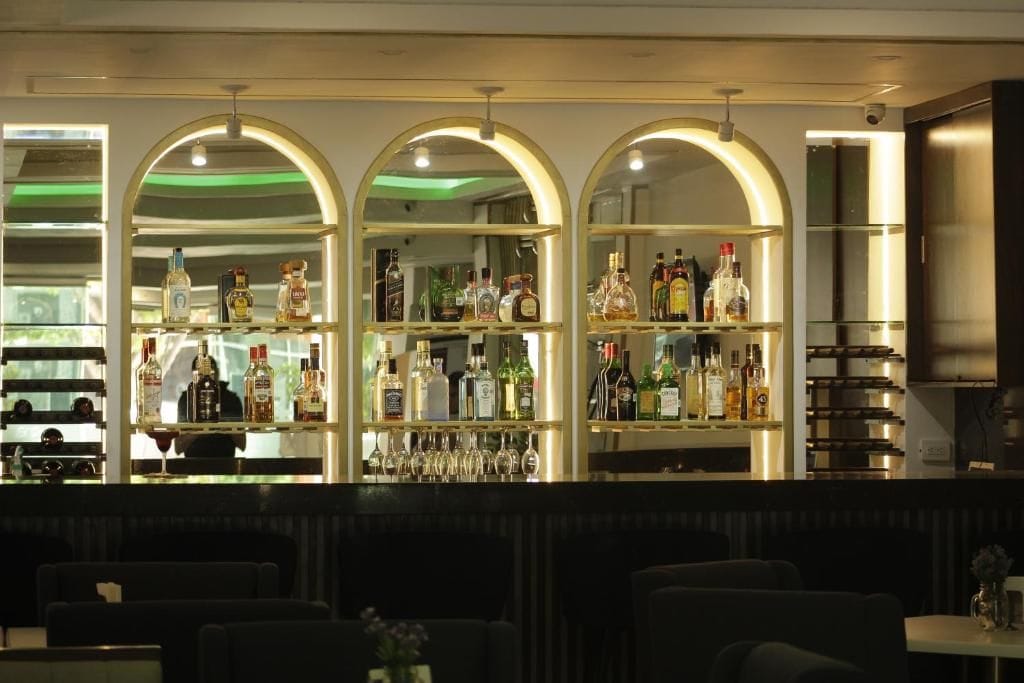 A bar area with multiple shelves of liquor bottles and glasses, and seating in the foreground.