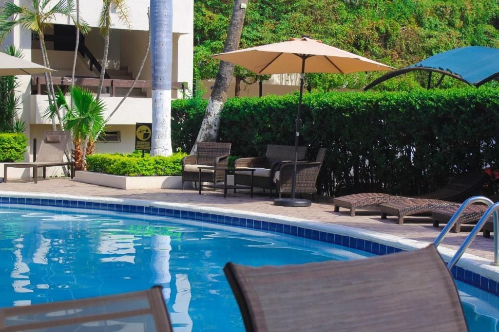 Una piscina con sillones de descanso y sombrillas, rodeada de vegetación y un edificio.