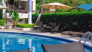 Una piscina con sillones de descanso y sombrillas, rodeada de vegetación y un edificio.
