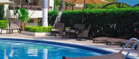 A swimming pool with lounge chairs and umbrellas, surrounded by greenery and a building.
