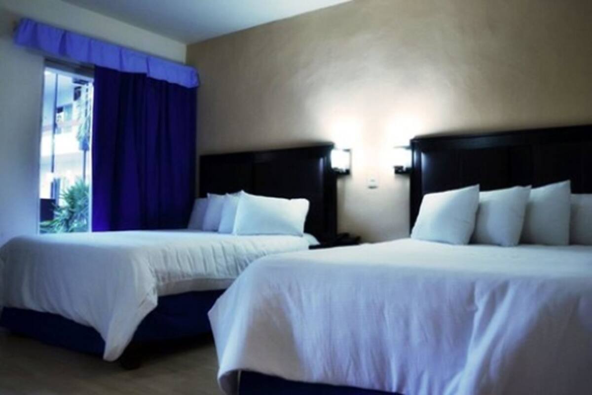 Two beds with white linens, a dark headboard, and a window with blue curtains.
