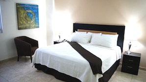 A neatly made bed with white and brown bedding, a bedside table with a lamp, a chair, and a painting on the wall. - Hotel El Dorado (Los Mochis)
