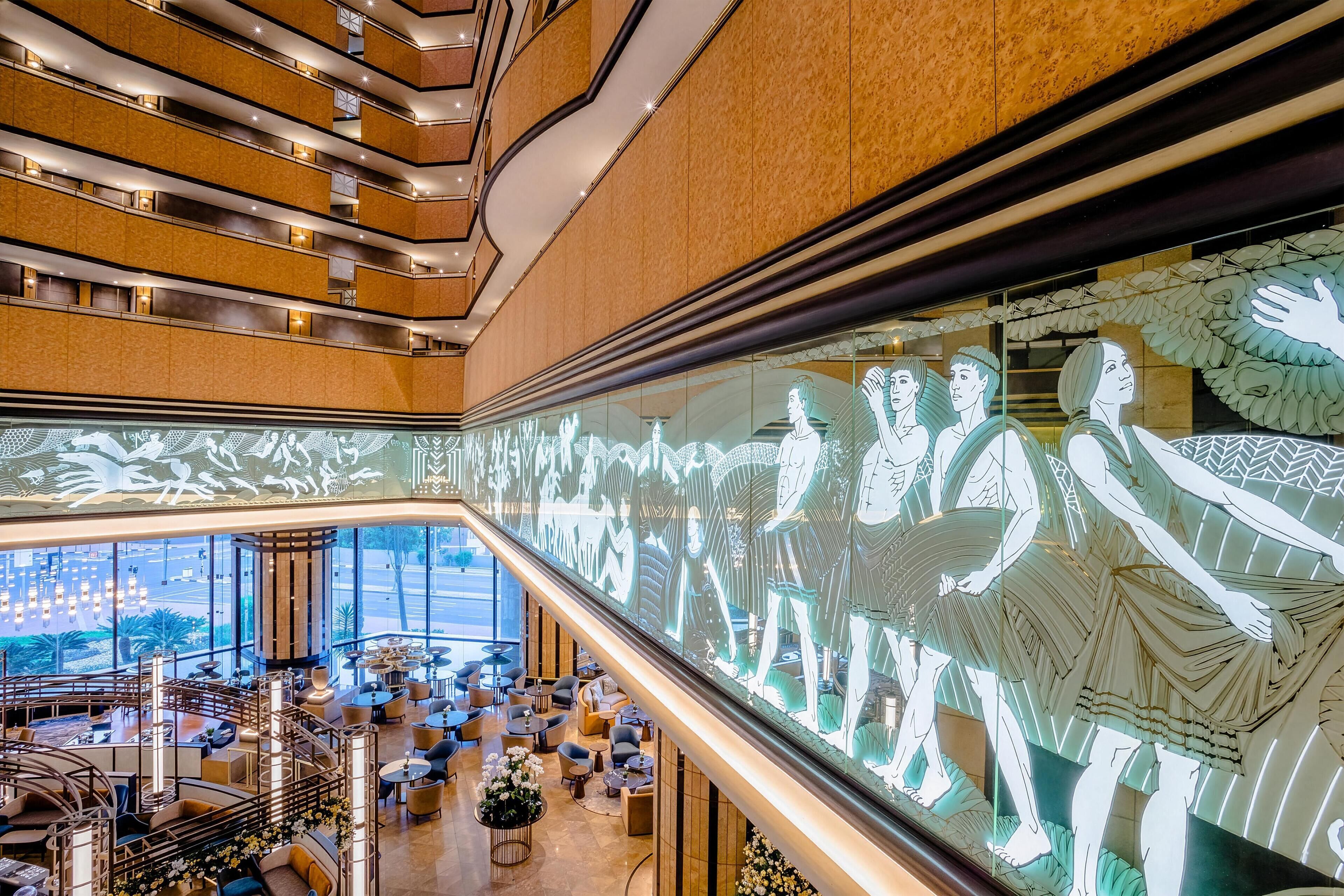 A modern hotel lobby with a large digital art display, a central seating area, and a view of the cityscape through large windows.