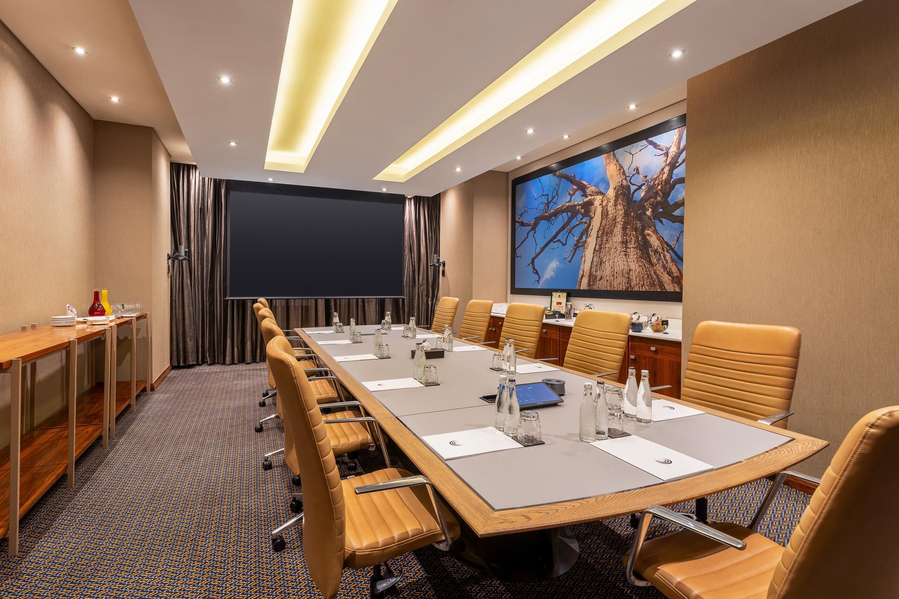 A conference room with a long table, chairs, a projector screen, and a large wall-mounted screen displaying a tree.