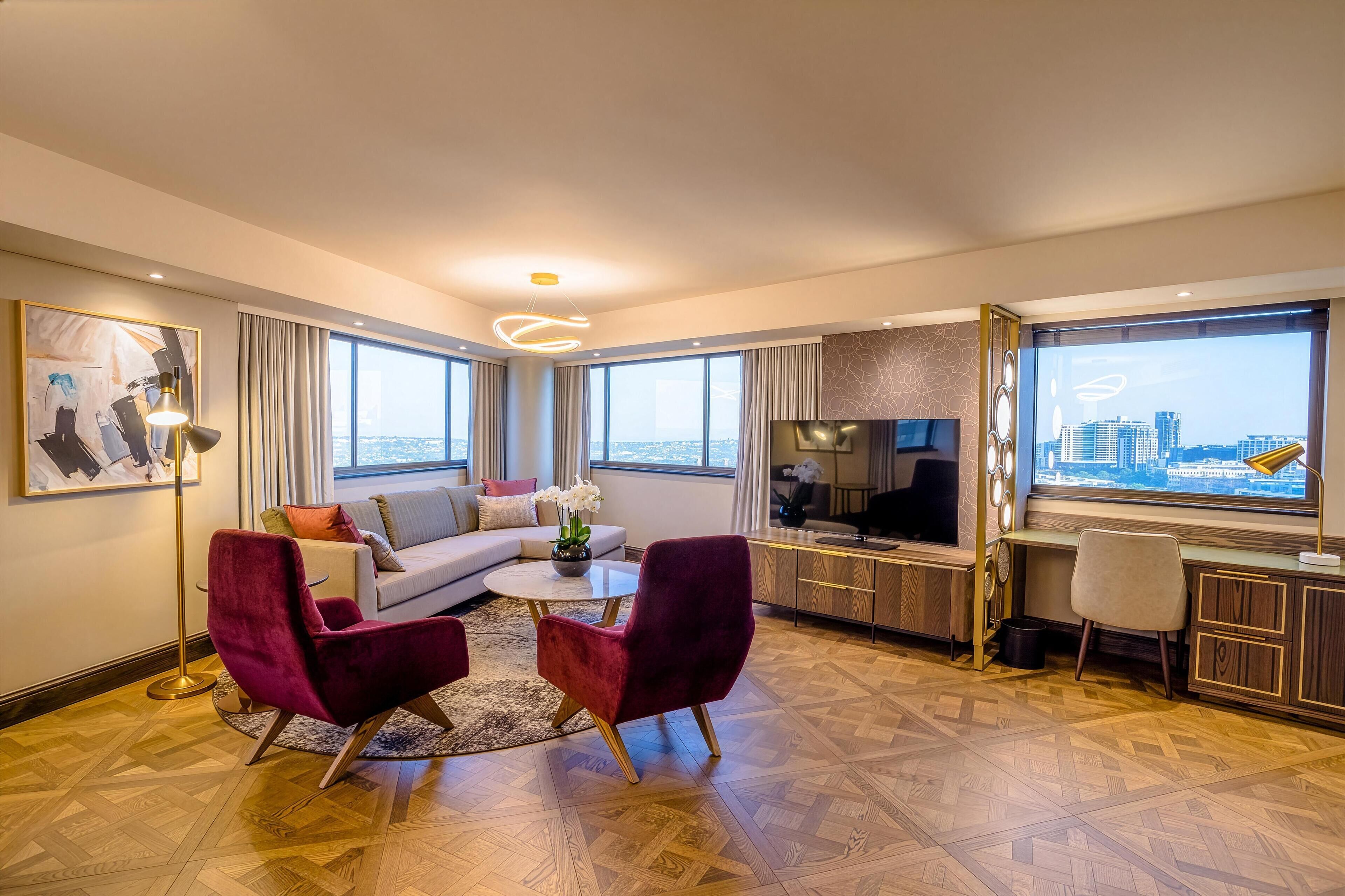 Una sala moderna con un sofá, dos sillones rojos, una mesa de centro, un televisor y un escritorio con una computadora. Amplias ventanas ofrecen vista a la ciudad.