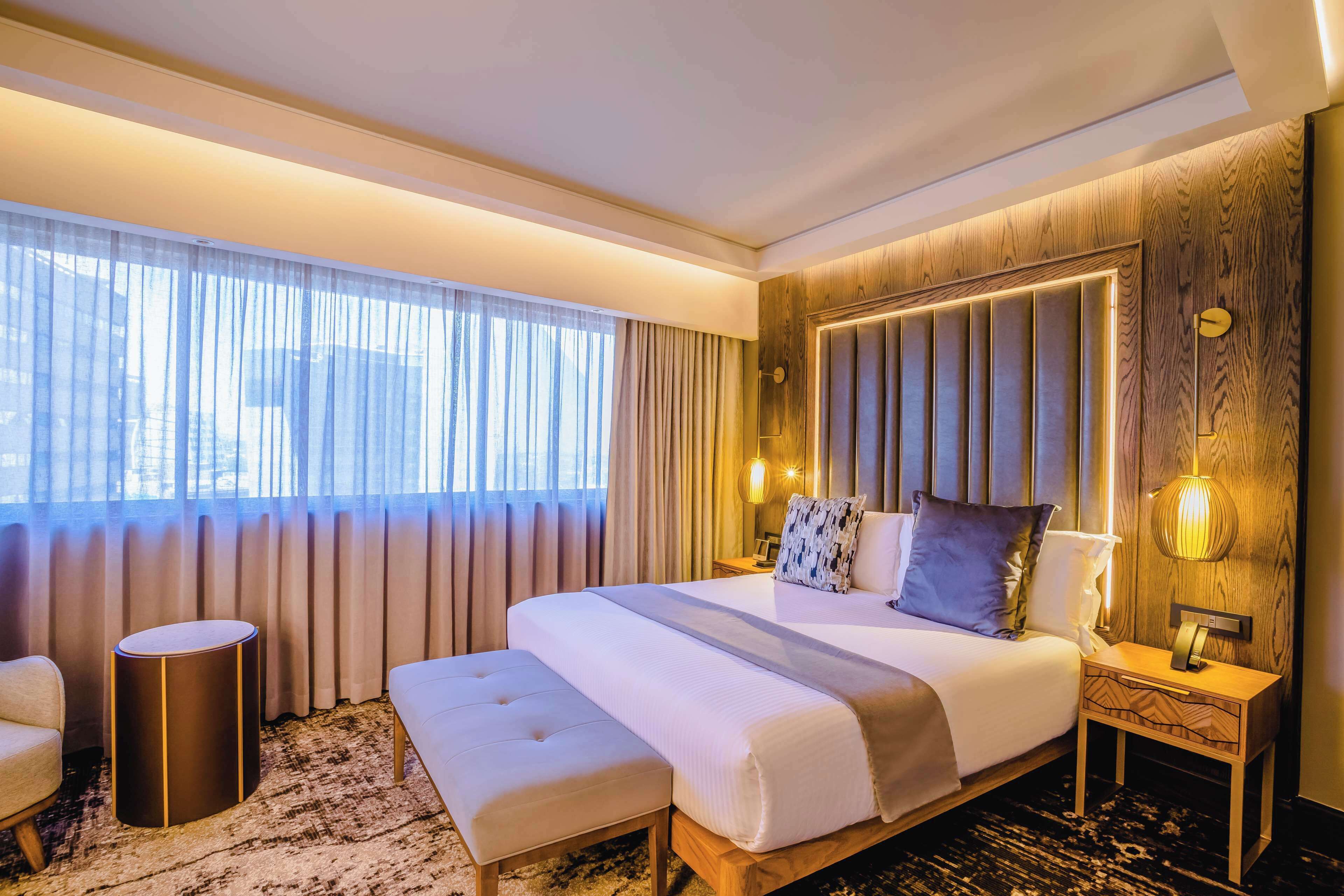 A hotel room with a large bed, a bedside table, a round side table, a bench, and a large window with curtains.