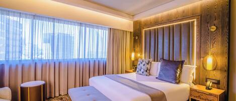 A hotel room with a large bed, a bedside table, a round side table, a bench, and a large window with curtains.