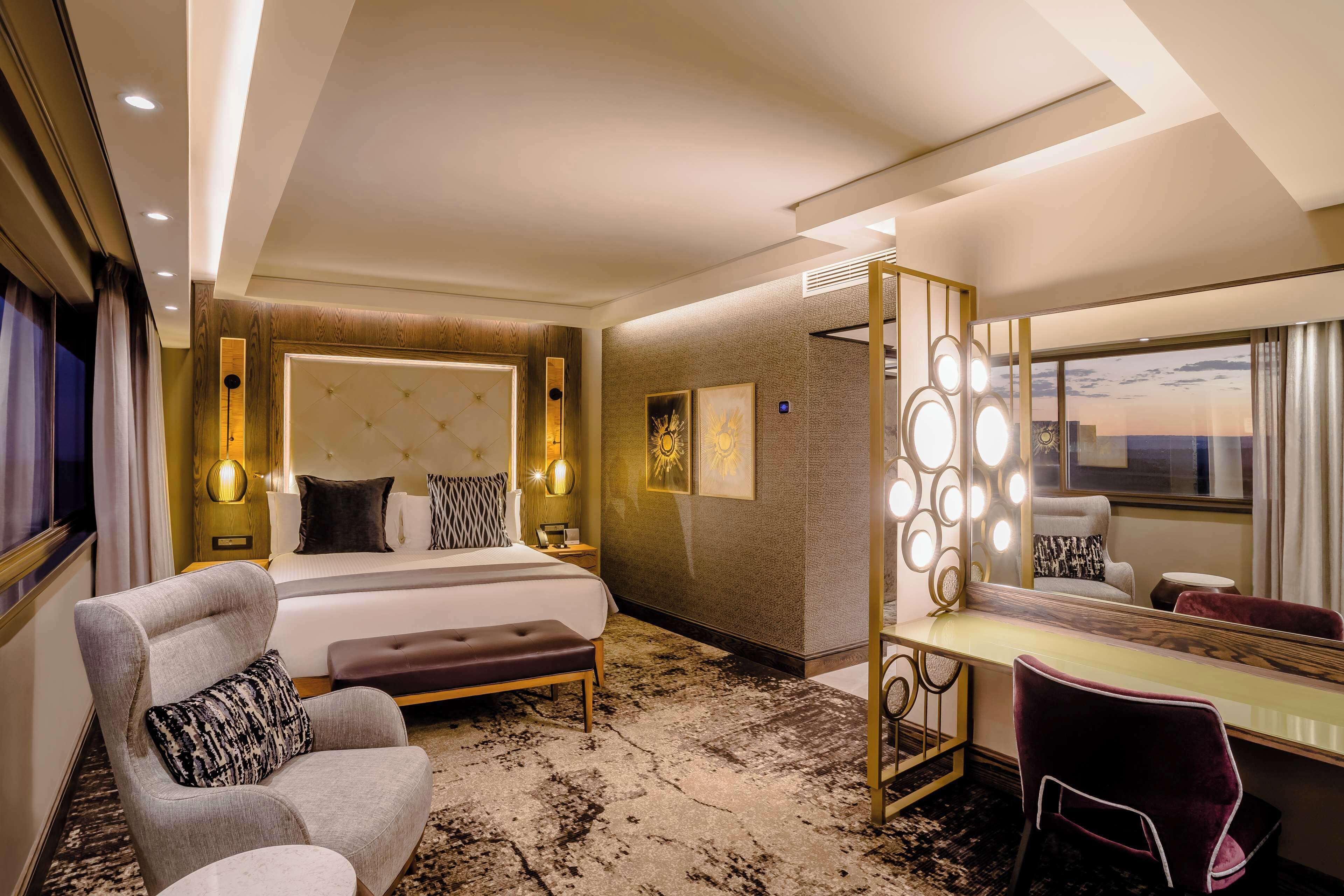 A hotel room with a large bed, a vanity with a mirror, a seating area, and a view of the sky.