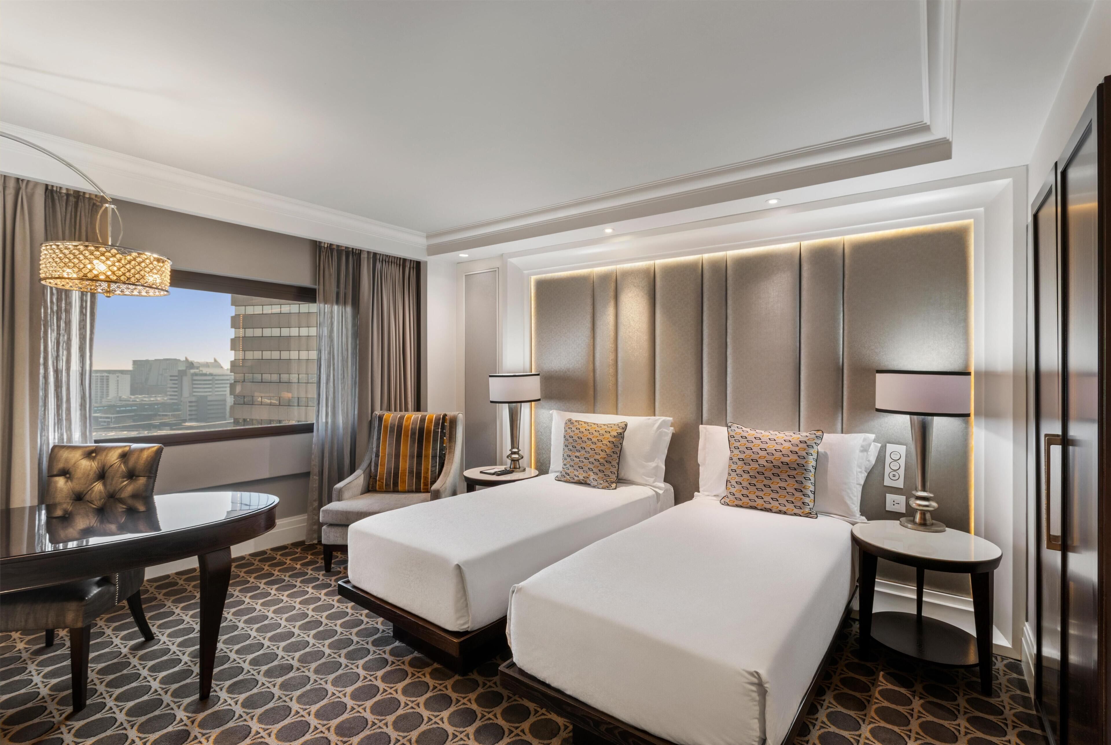 Habitación de hotel con dos camas, una mesa de comedor, una silla, una lámpara y vistas a la ciudad.