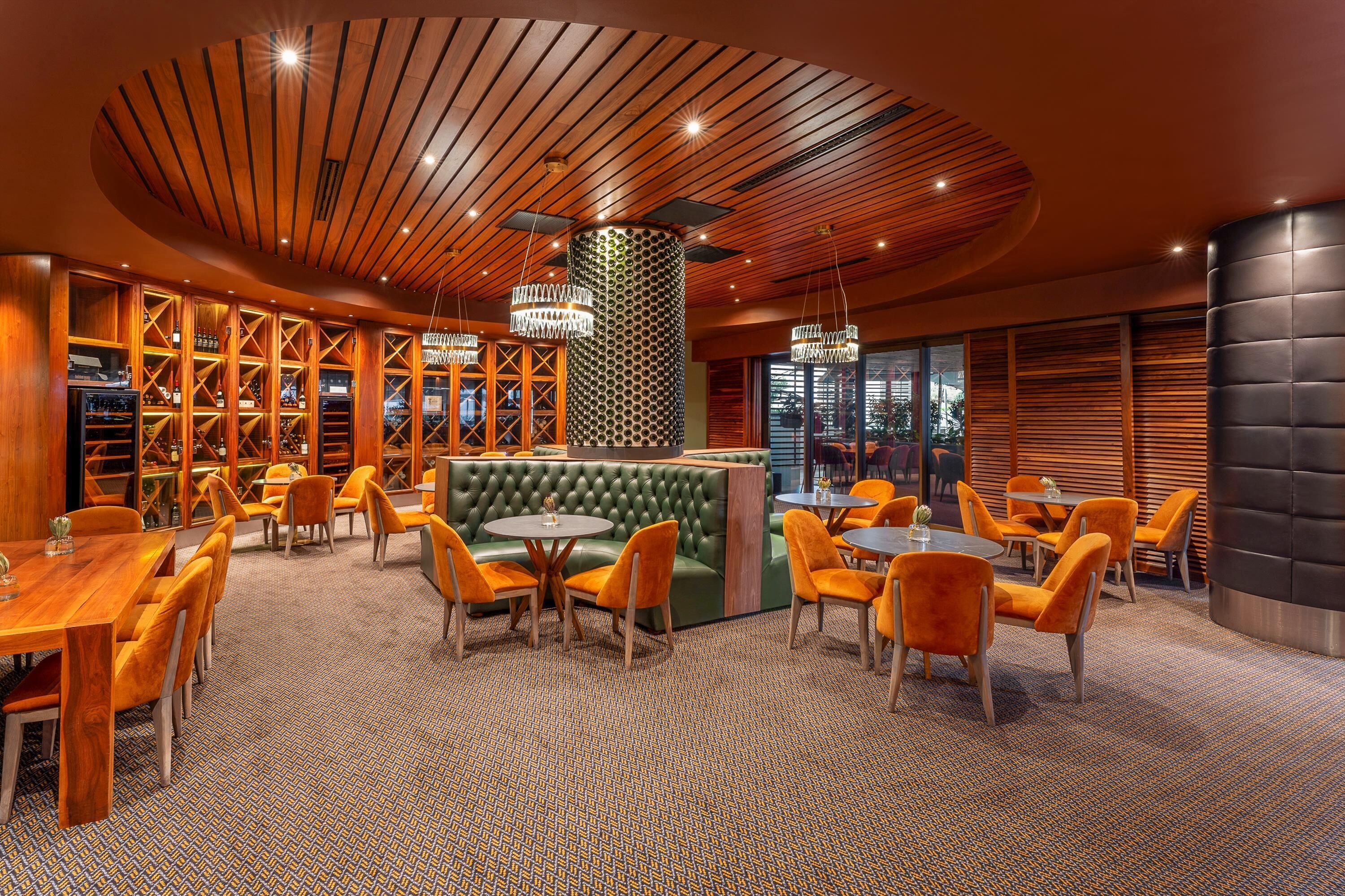 A modern bar area with wooden shelves, a green cushioned seating area, and orange chairs.