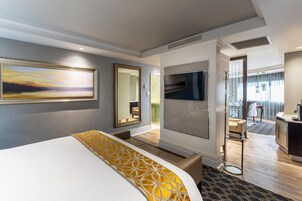 A hotel room with a large bed, a painting on the wall, a mirror, a television, and a view of the hallway.