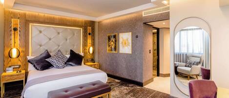 Une chambre d’hôtel moderne dotée d’un grand lit, d’une banquette, d’une table de chevet, d’un miroir et d’un coin salon avec un fauteuil.