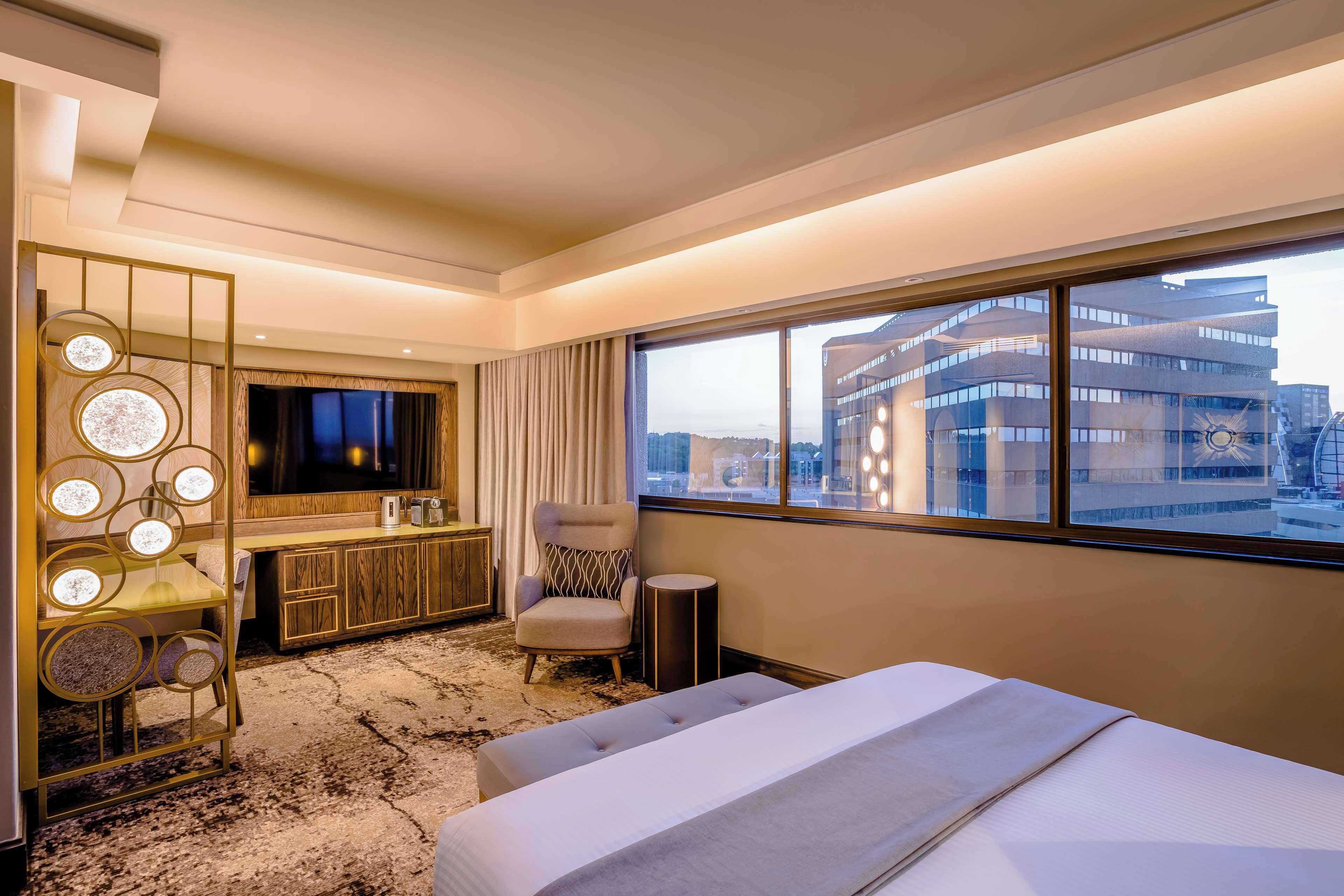 A hotel room with a large bed, a desk with a TV, a chair, and a view of a building.