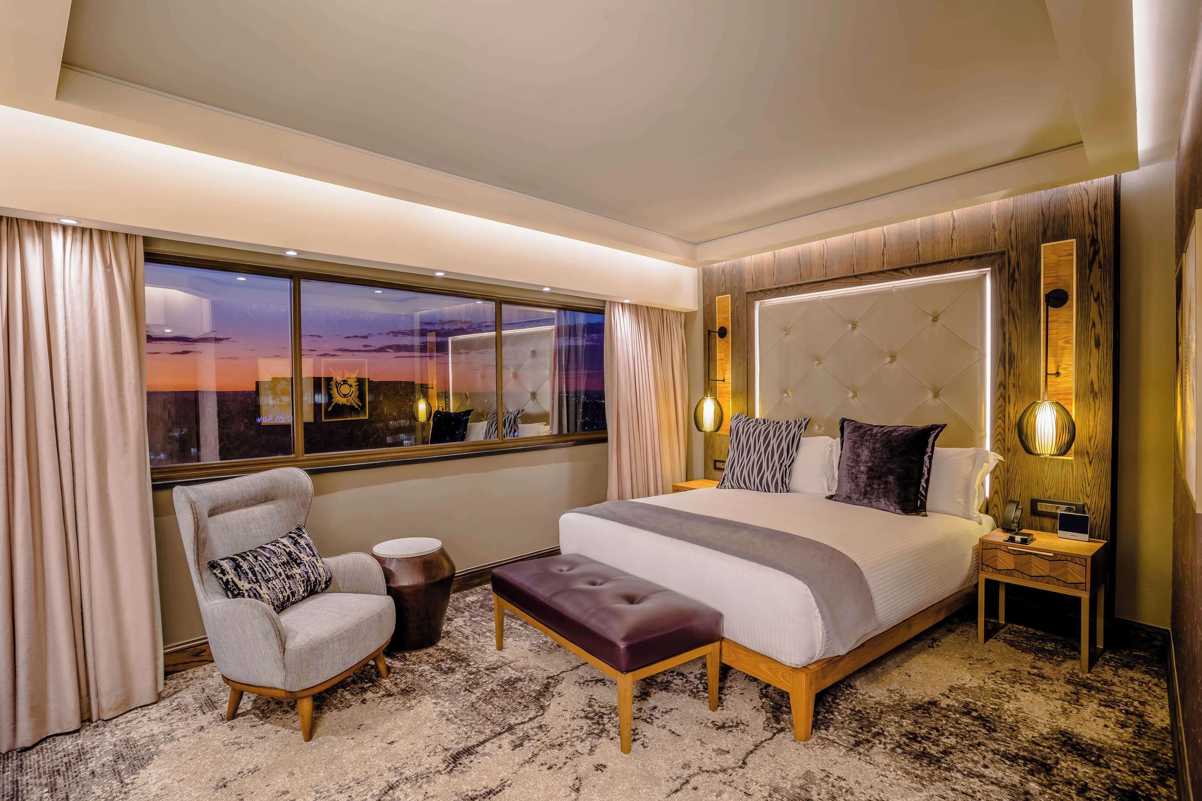 Habitación de hotel moderna con una cama grande, una zona de estar con una silla y reposapiés, una mesita de noche y vistas a la puesta de sol a través de la ventana.