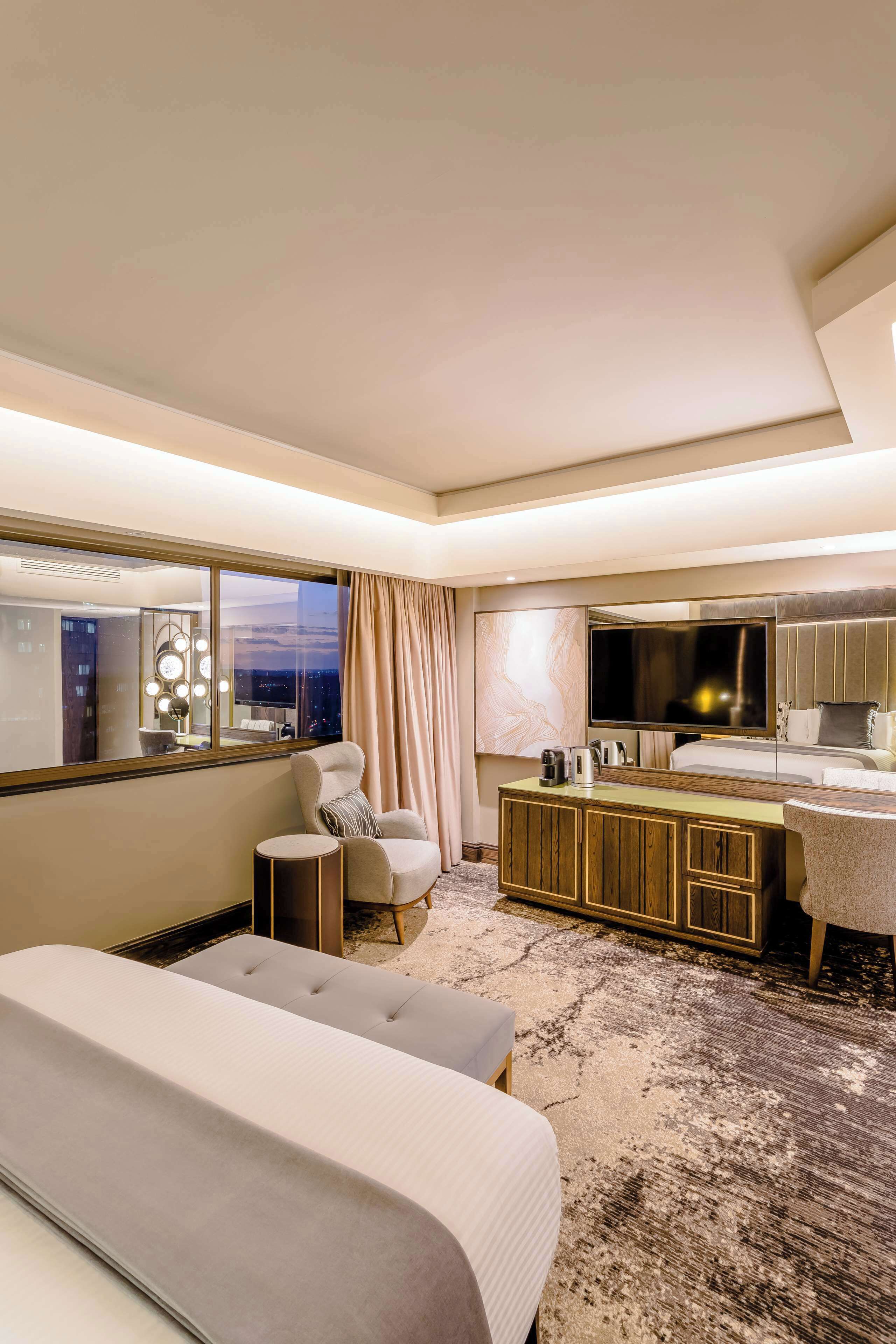 Una habitación de hotel moderna con una cama grande, una zona de estar con sofá y sillón, una mesita, un televisor y un minibar.