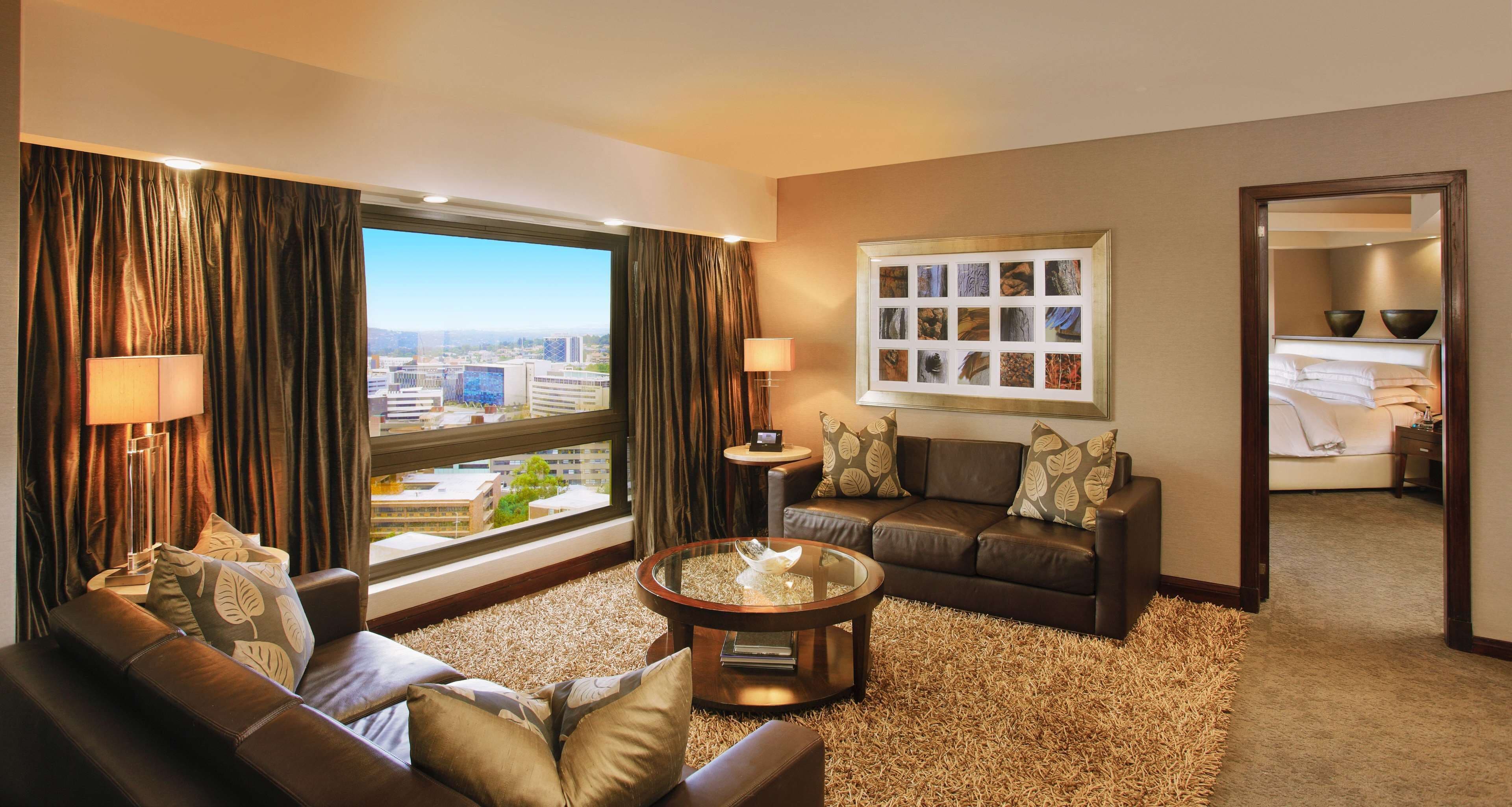 A modern hotel room with a leather sofa, a round coffee table, and a large window offering a city view.