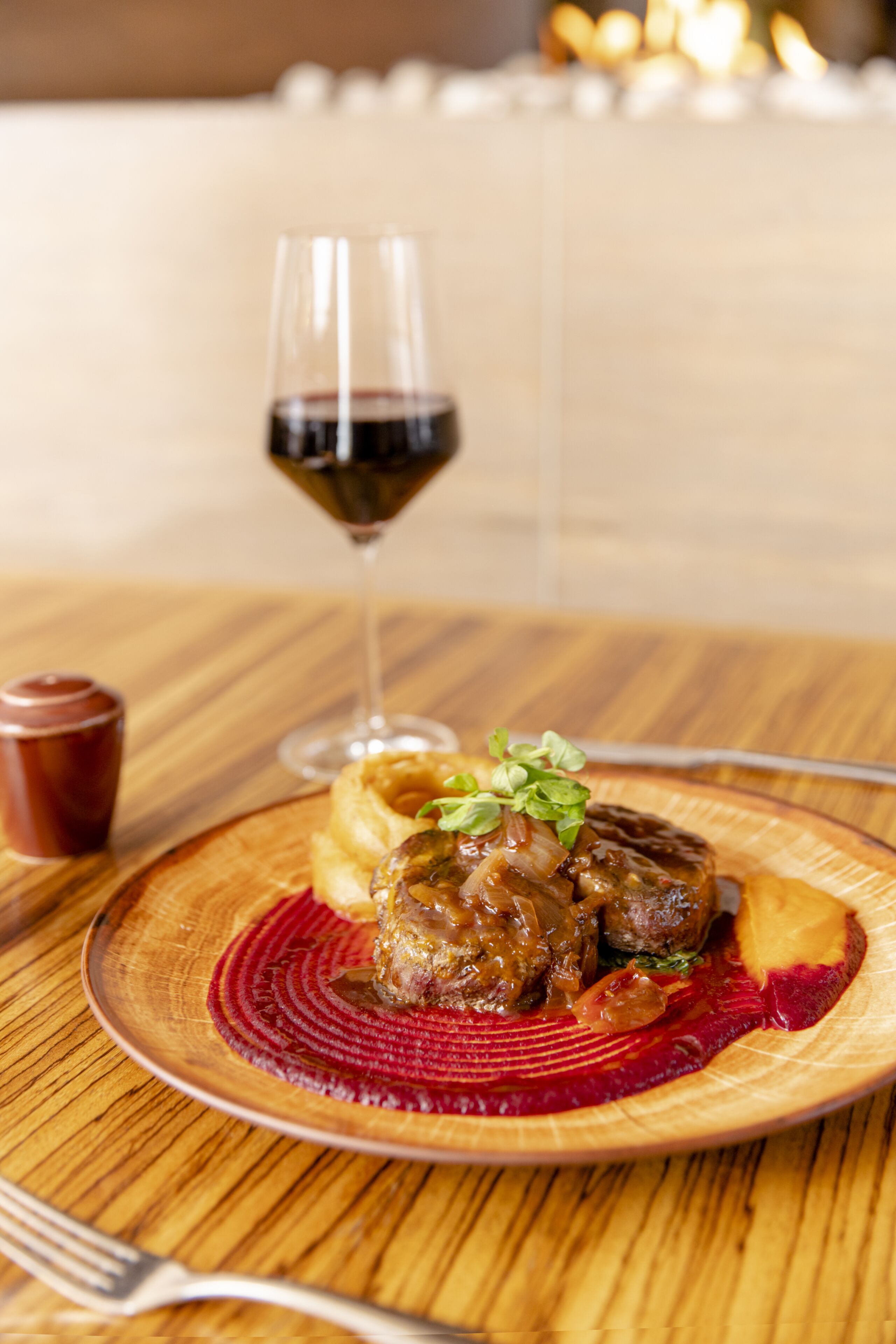 A wooden table with a plate of food, a glass of red wine, and a small cup.