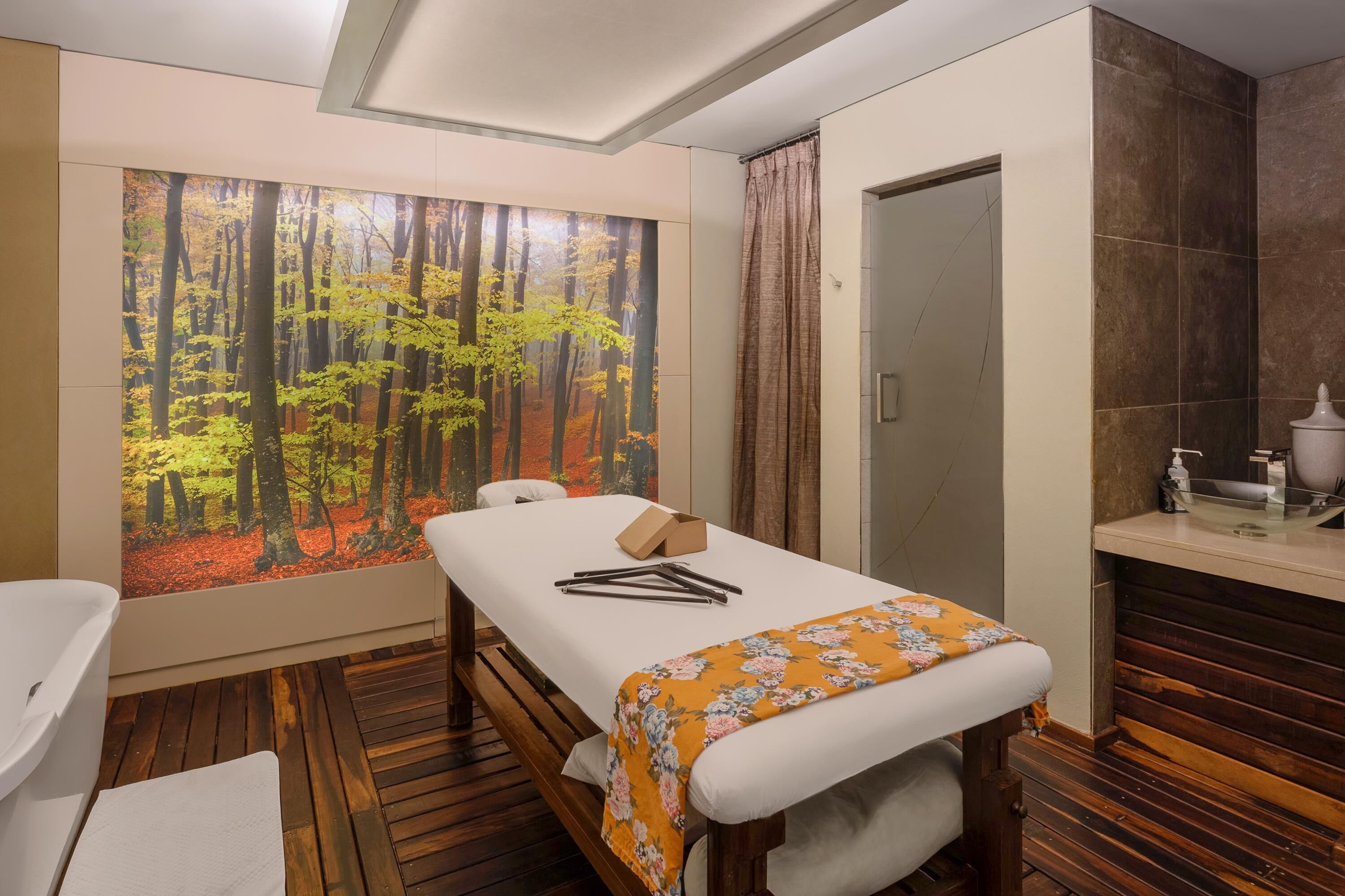 Una habitación con piso de madera, cama con colchón blanco y colcha naranja con estampado floral, un banco de madera, un ventanal con un mural de un bosque y un baño con lavamanos y espejo.