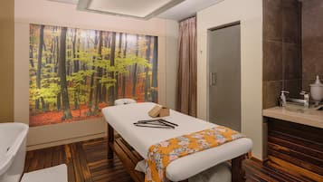 Una habitación con piso de madera, cama con colchón blanco y colcha naranja con estampado floral, un banco de madera, un ventanal con un mural de un bosque y un baño con lavamanos y espejo.