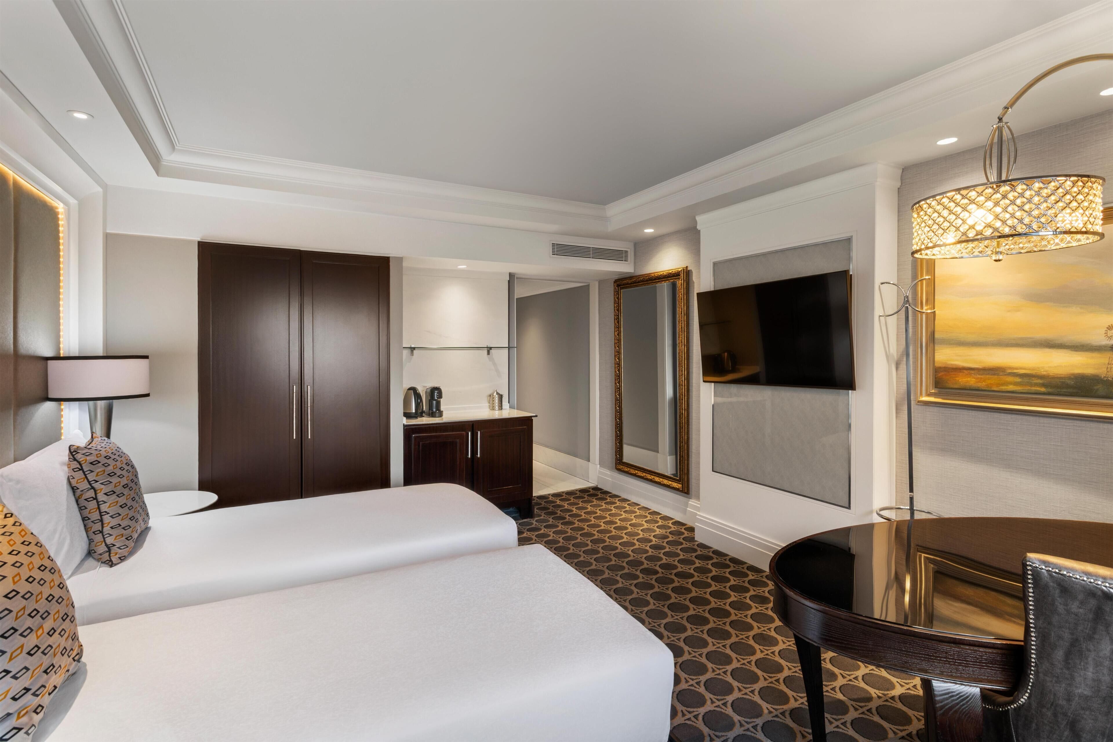 A hotel room with a bed, a desk, a chair, a large mirror, and a painting of a sunset.