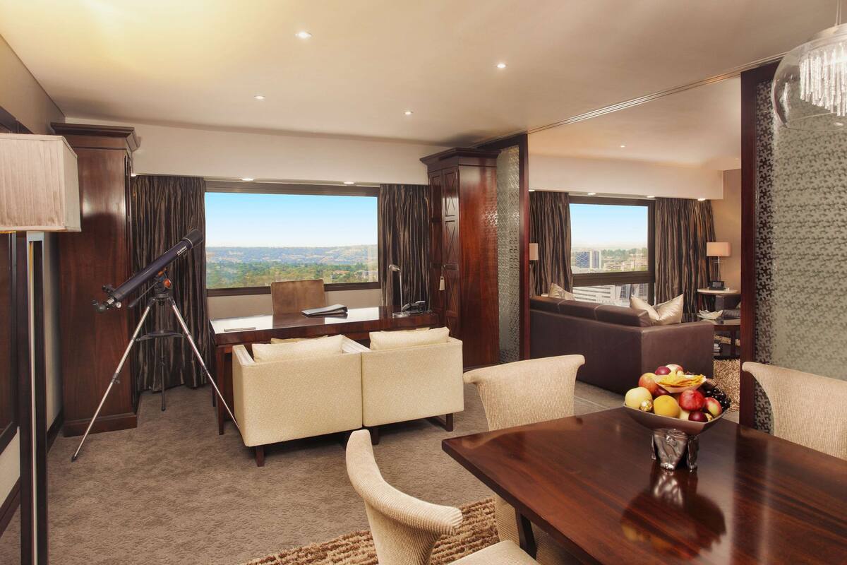 Ein Hotelzimmer mit großem Fenster und Blick auf die Stadt, einem Holztisch, einer Couch und einem Esstisch mit Obst.