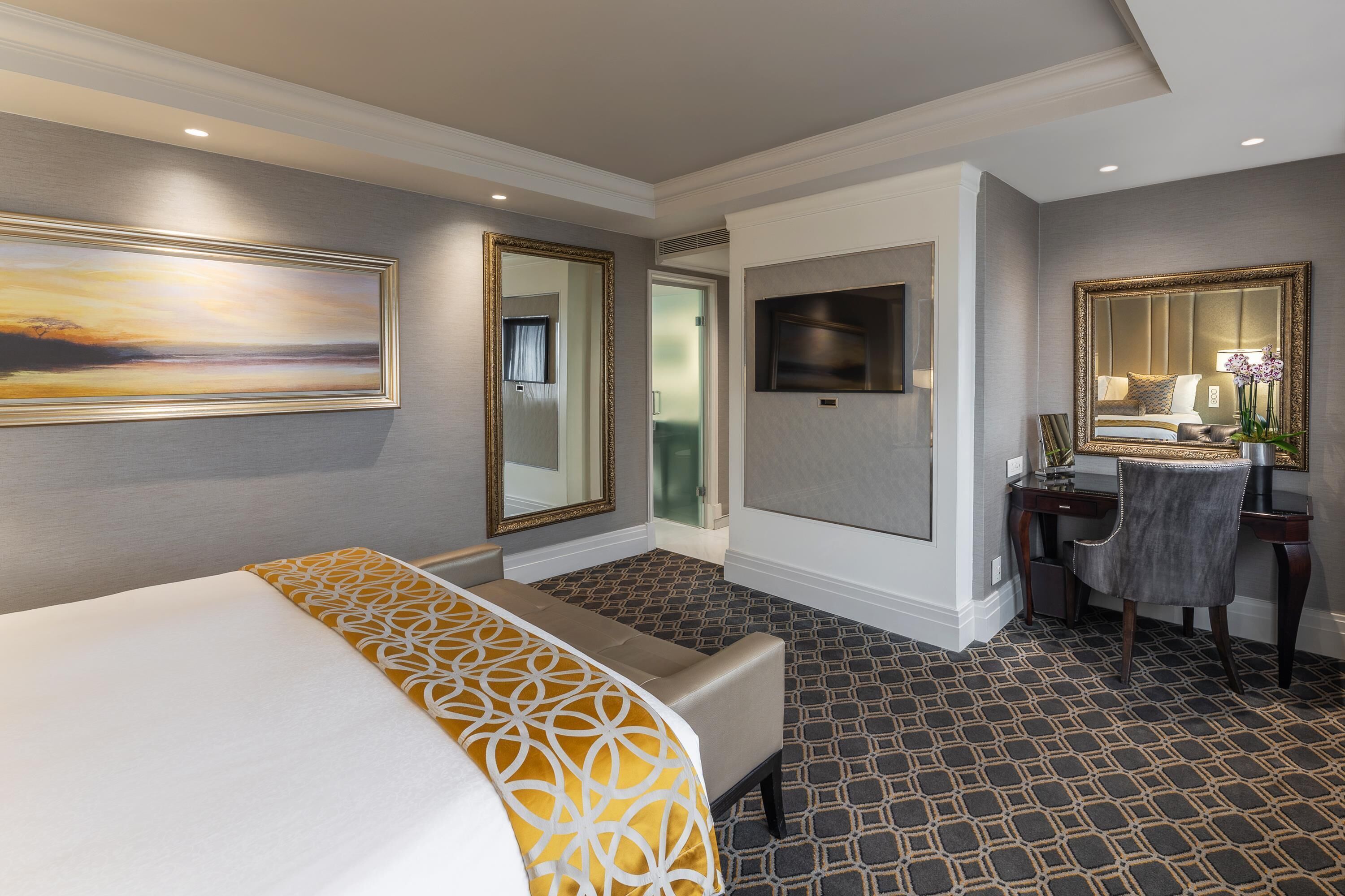 A hotel room with a bed, a desk, a chair, a mirror, and a painting on the wall.