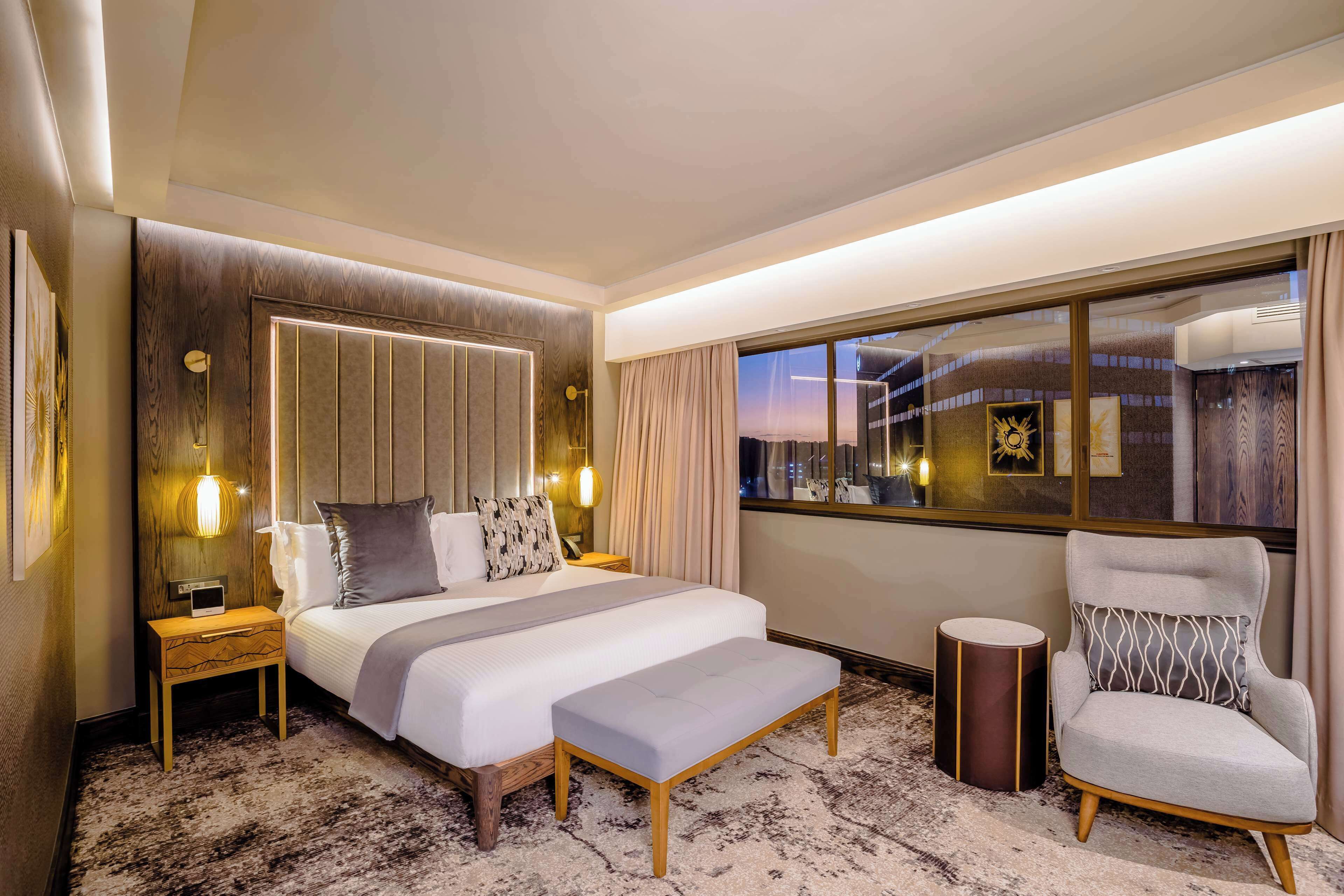 A hotel room with a large bed, bedside tables, a chair, a small table, and a large window with a view of the city.