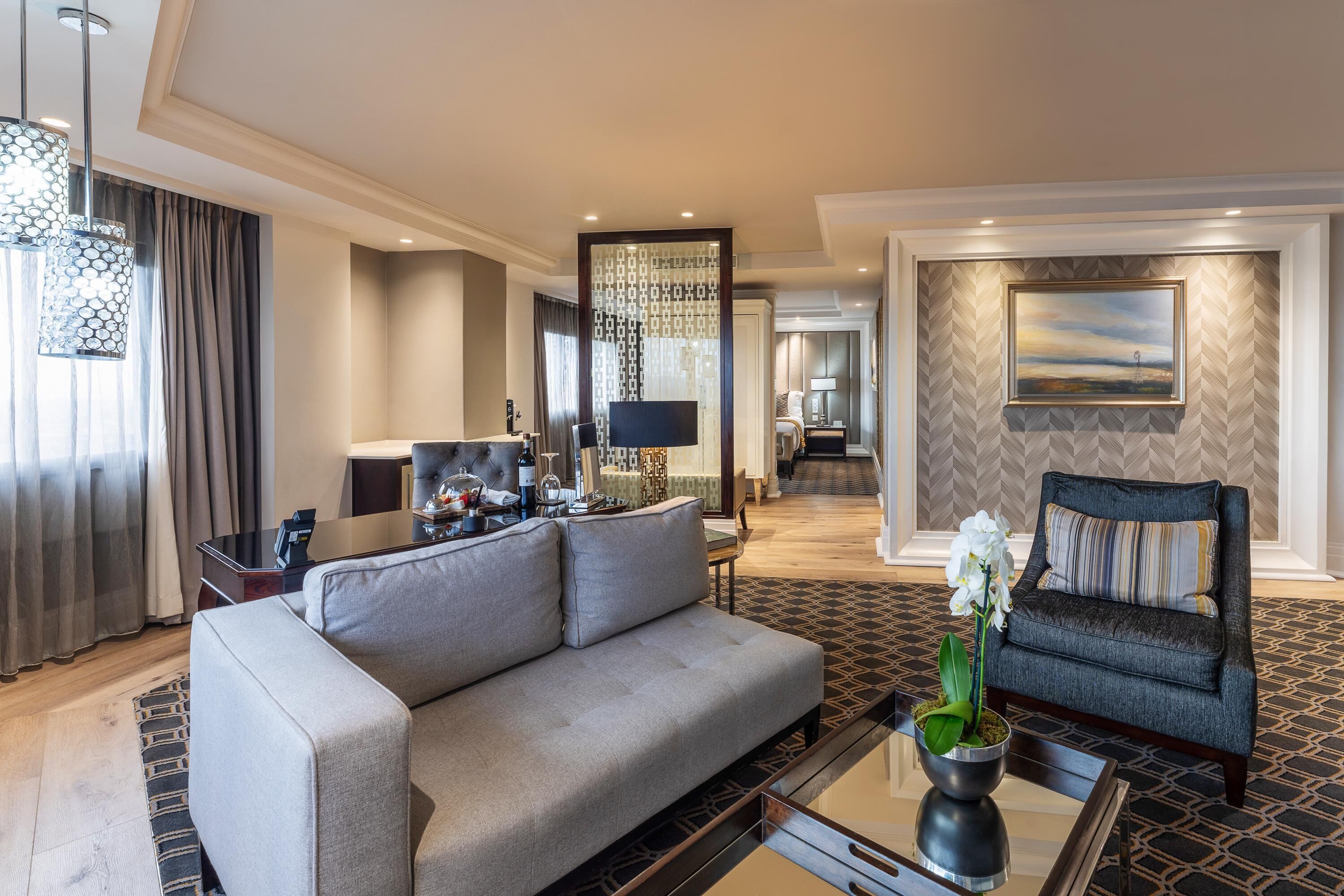 A modern hotel room with a sofa, a dining table, and a coffee table. There is a chandelier, a painting on the wall, and a view of the outside.
