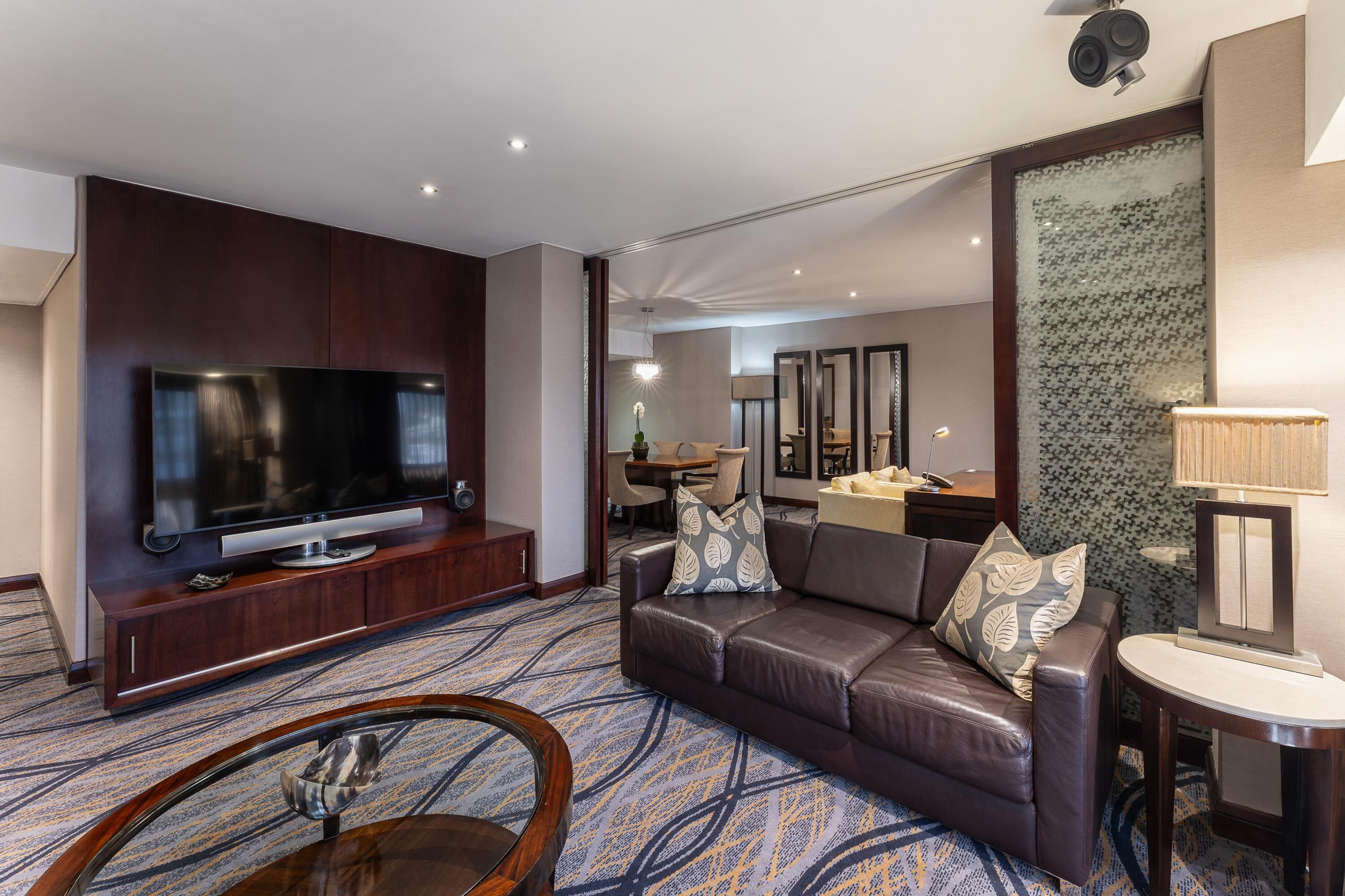 Una habitación de hotel moderna con televisor de pantalla plana, sofá de cuero, alfombra estampada y una zona de comedor al fondo.
