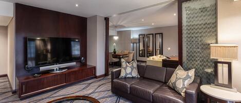 A modern hotel room with a flat-screen TV, leather sofa, patterned carpet, and a dining area in the background.