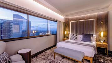 A modern hotel room with a large bed, a bench, a round side table, and a view of the cityscape through large windows.