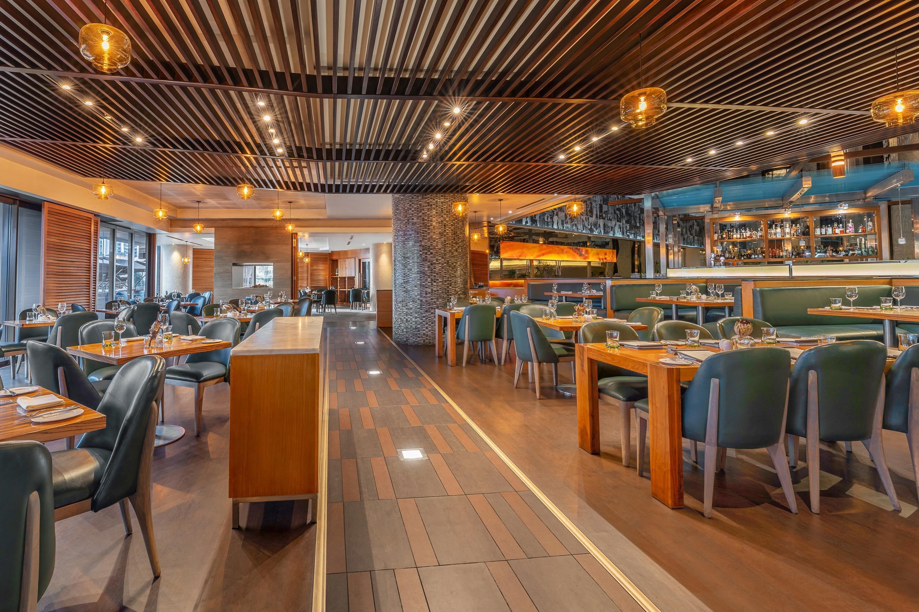 A modern restaurant interior with wooden flooring, green upholstered booths, and a bar area with a visible liquor display.
