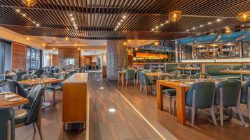 A modern restaurant interior with wooden flooring, green upholstered booths, and a bar area with a visible liquor display.