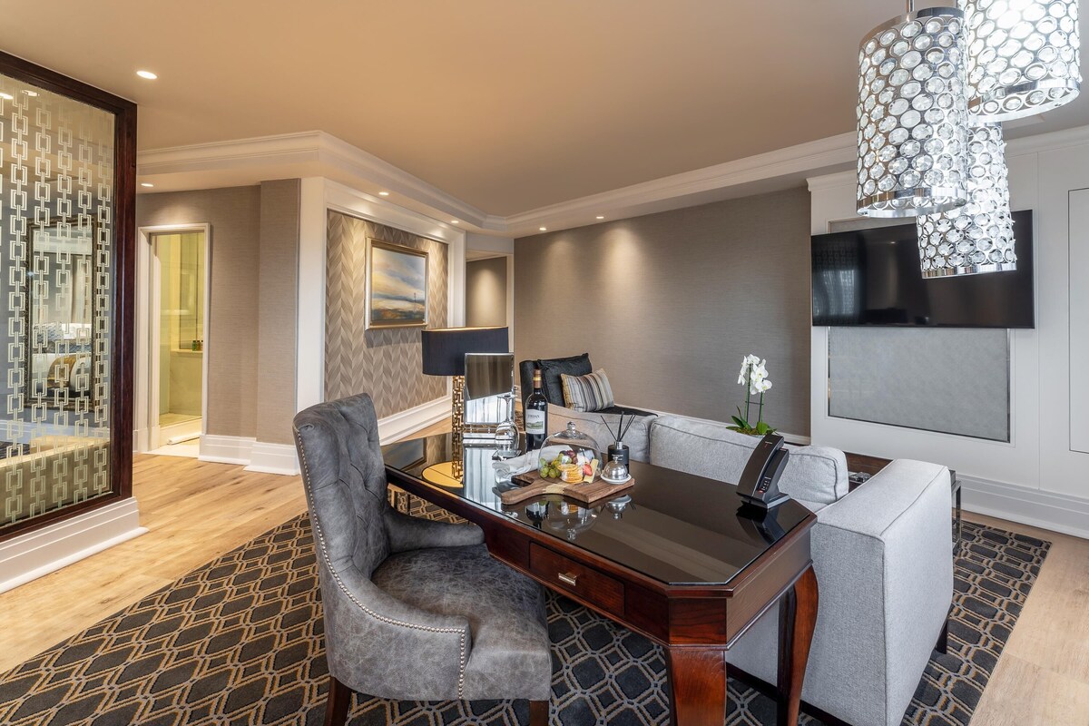 A modern hotel room with a dining area, a sofa, a desk with a lamp, and a decorative chandelier.