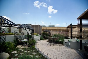 Un patio al aire libre con arreglos de asientos, incluyendo mesas y sillas, rodeado de plantas y un sendero de ladrillo.