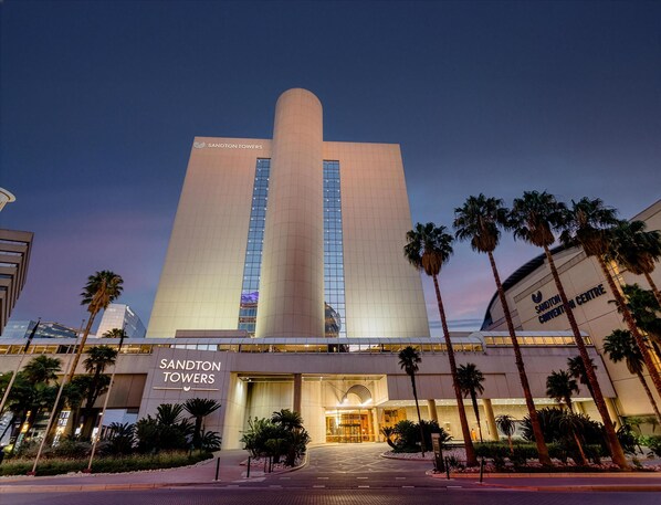 A modern hotel with a prominent central tower, a well-lit entrance, and palm trees in the foreground. - Sandton Sun & Towers (Johannesburg)