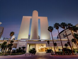 Un hotel moderno con una imponente torre central, una entrada bien iluminada y palmeras en primer plano.