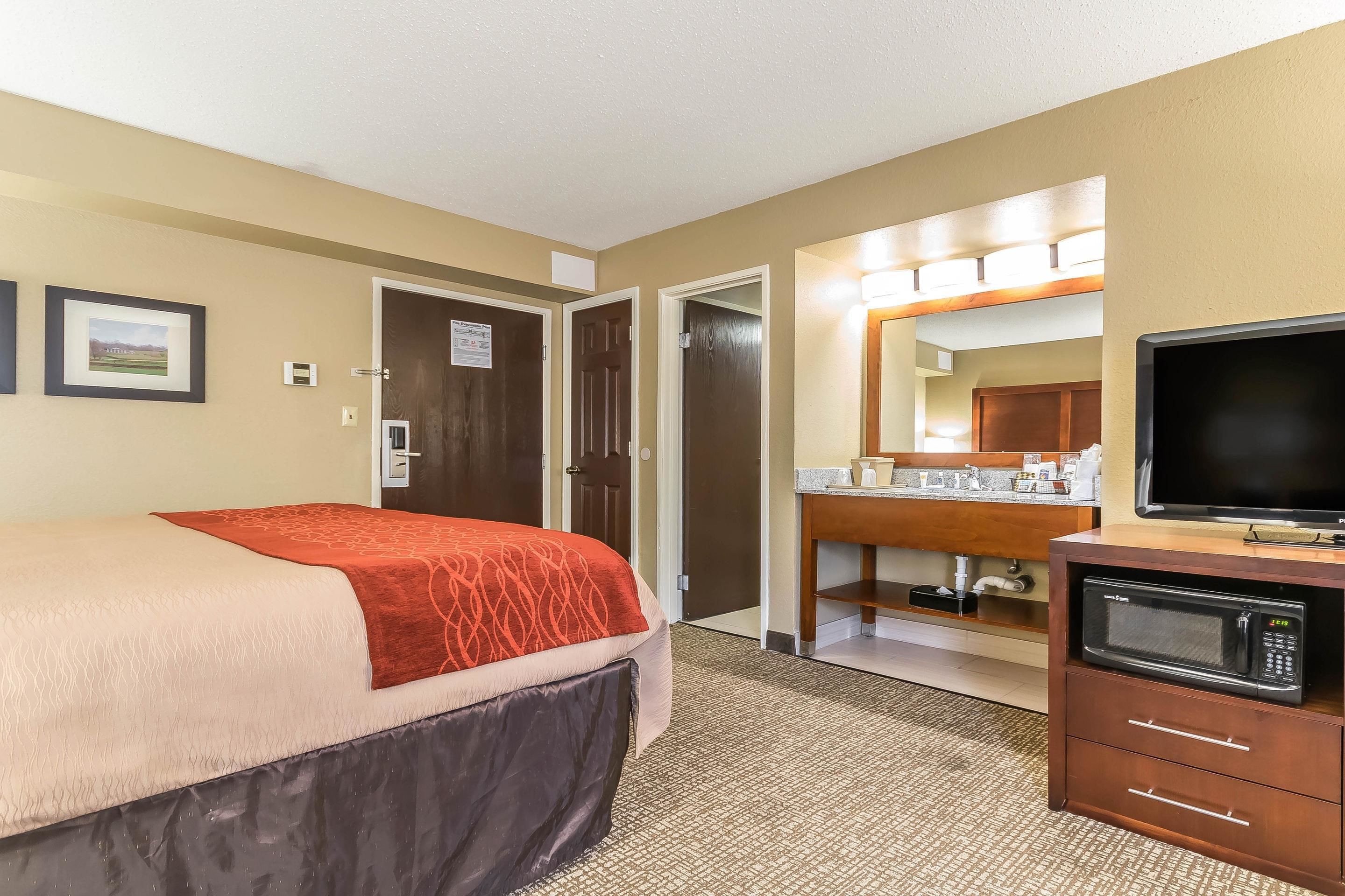 Habitación de hotel con cama, escritorio con microondas, televisor y baño visible a través de una puerta abierta.