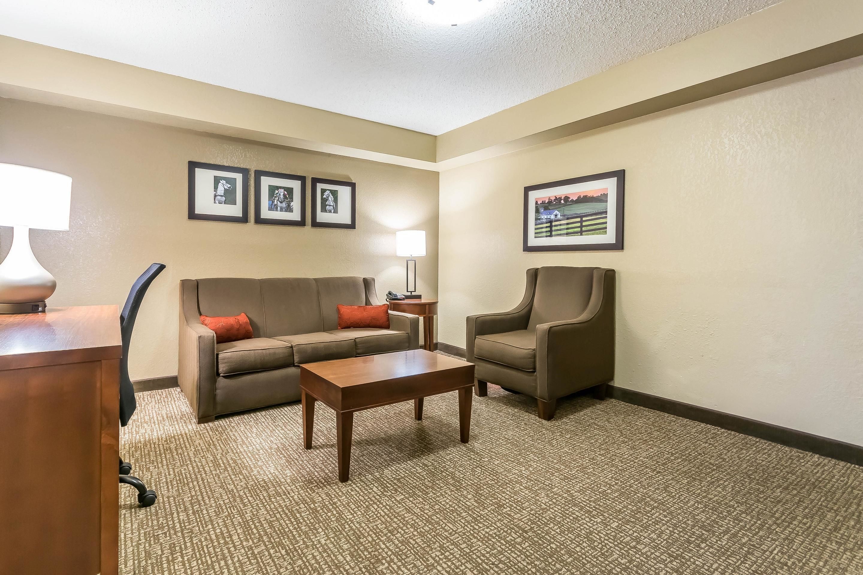 Habitación de hotel con sofá, sillón, mesa de centro y escritorio con lámpara. En la pared hay cuadros enmarcados y el suelo está alfombrado.