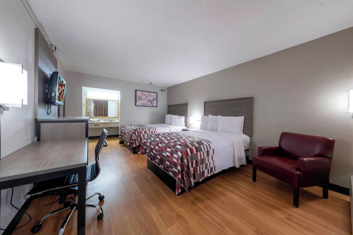 A hotel room with a wooden floor, a desk with a chair, two beds with patterned bedspreads, a red leather armchair, and a flat-screen TV mounted on the wall.