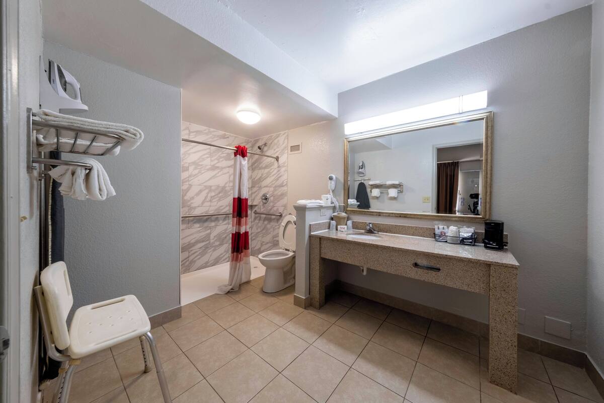 A bathroom with a shower, toilet, and sink. There is a chair, a towel rack, and a mirror.