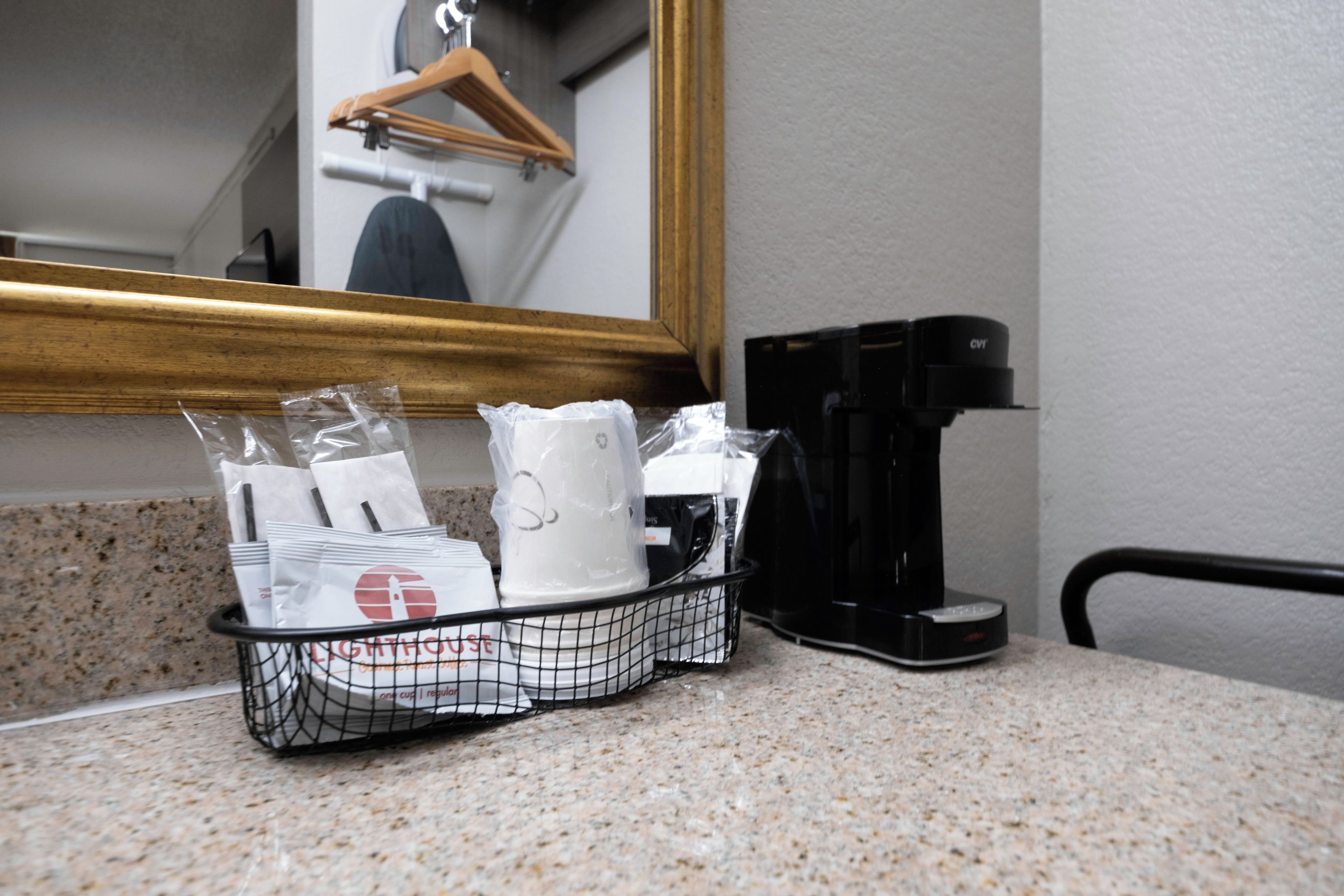 Un comptoir dans une chambre d’hôtel, avec une machine à café, un panier contenant des articles de toilette et un miroir agrémenté d’un cintre en bois.