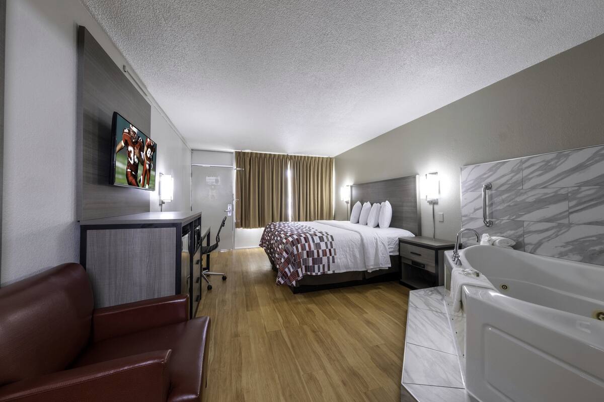 A modern hotel room with a bed, a desk, a chair, a bathtub, and a TV on the wall.